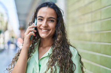Genç, güzel, İspanyol bir kadın gülümsüyor ve caddede akıllı telefondan konuşuyor.