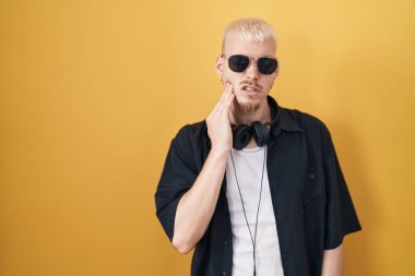 Beyaz güneş gözlüğü takan genç bir adam diş ağrısı ya da diş ağrısı yüzünden sarı arka planda durup ağız ve acı dolu bir ifadeyle dokunuyor. dişçi 