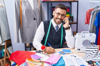 Genç İspanyol terzi terzide akıllı telefondan konuşuyor. Terzide defter üzerinde yazıyor.