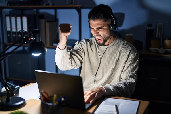 Young handsome man working using computer laptop at night angry and mad raising fist frustrated and furious while shouting with anger. rage and aggressive concept.