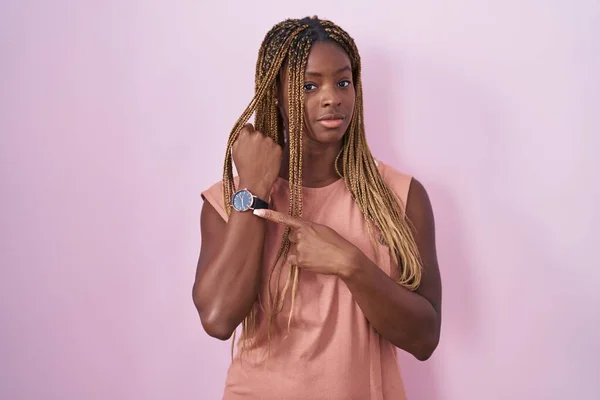 African american woman with braided hair standing over pink background in hurry pointing to watch time, impatience, looking at the camera with relaxed expression