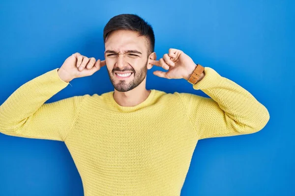 青い背景の上に立つヒスパニック系の男は 大きな音楽のノイズのためのいらいらした表情で指で耳を覆う 聴覚障害の概念 — ストック写真