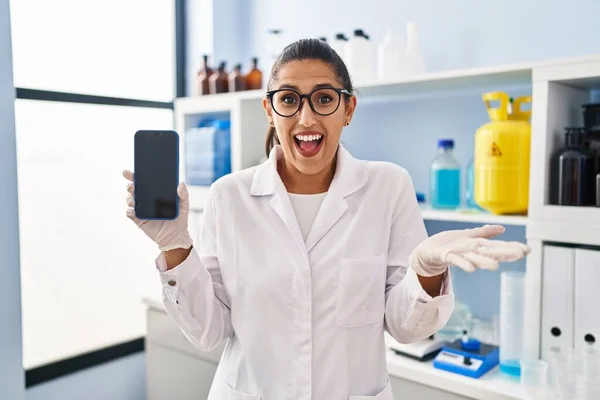 幸せな笑顔で達成を祝うスマートフォンで科学研究所で働く若いヒスパニック系女性と手を挙げて勝者の式 — ストック写真
