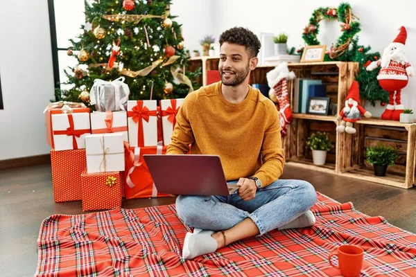 顔に笑みを浮かべて横に見てクリスマスツリーに座ってノートパソコンを使用してアラブの若い男は 自然な表現 自信を持って — ストック写真