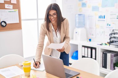 Young beautiful hispanic woman business worker reading document writing on notebook at office