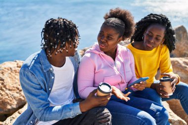 African american friends using smartphone and drinking coffee sitting on rock at seaside
