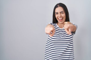 Çizgili tişört giyen genç esmer kadın seni ve kamerayı işaret ediyor, gülümsüyor olumlu ve neşeli. 