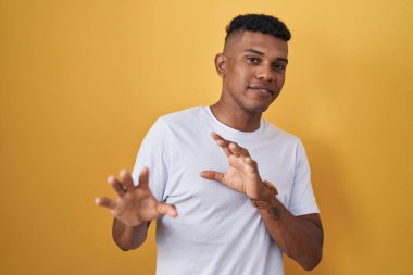 Young hispanic man standing over yellow background disgusted expression, displeased and fearful doing disgust face because aversion reaction. 