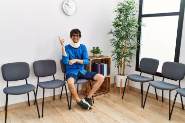 Young hispanic man sitting at doctor waiting room with neck injury smiling happy pointing with hand and finger to the side 
