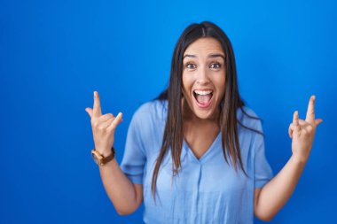 Young brunette woman standing over blue background shouting with crazy expression doing rock symbol with hands up. music star. heavy music concept. 