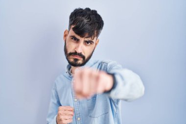 Mavi arka planda sakallı genç bir İspanyol dövüşmek için yumrukluyor, agresif ve öfkeli saldırı, tehdit ve şiddet 