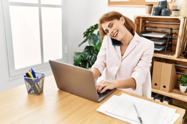 Ofiste çalışan genç beyaz iş kadını bilgisayarla akıllı telefondan konuşuyor..