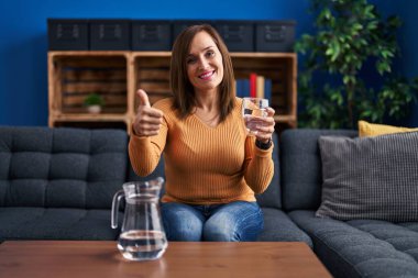 Middle age woman drinking glass of water smiling happy and positive, thumb up doing excellent and approval sign 