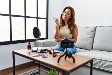 Middle age hispanic woman recording make up tutorial with smartphone at home smiling happy pointing with hand and finger to the side 