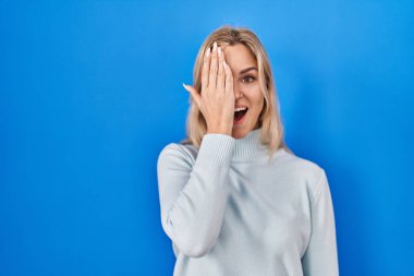 Mavi arka planda duran genç beyaz kadın bir gözünü eliyle kapatıyor, yüzünde kendine güvenen bir gülümseme ve sürpriz bir duygu var.. 