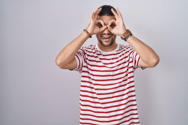 Hispanic man with long hair standing over isolated background doing ok gesture like binoculars sticking tongue out, eyes looking through fingers. crazy expression. 