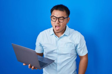 Chinese young man using computer laptop angry and mad screaming frustrated and furious, shouting with anger. rage and aggressive concept. 