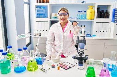 Young hispanic woman working at scientist laboratory doing make up looking positive and happy standing and smiling with a confident smile showing teeth 