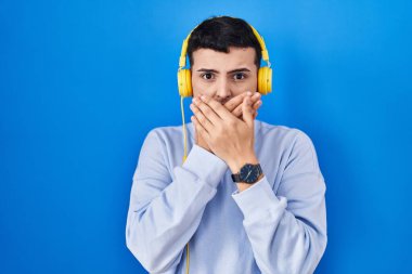 Non binary person listening to music using headphones shocked covering mouth with hands for mistake. secret concept. 
