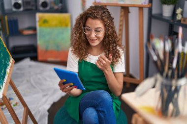 Beyaz kadın ressam, resim stüdyosunda oturmuş, mutlu ve pozitif gülümseyen tabletle video çağrısı yapıyor, başparmağıyla mükemmel ve onay işareti yapıyor. 