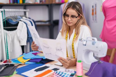 Young blonde woman tailor looking clothing design at clothing shop