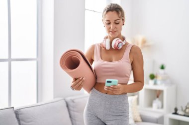 Young blonde woman using smartphone holding yoga mat at home