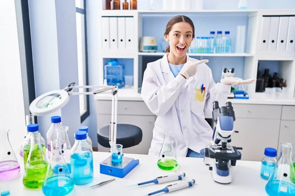 Young brunette woman working at scientist laboratory amazed and smiling to the camera while presenting with hand and pointing with finger.
