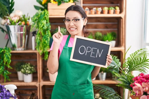 Hispanic young woman working at florist holding open sign smiling with an idea or question pointing finger with happy face, number one