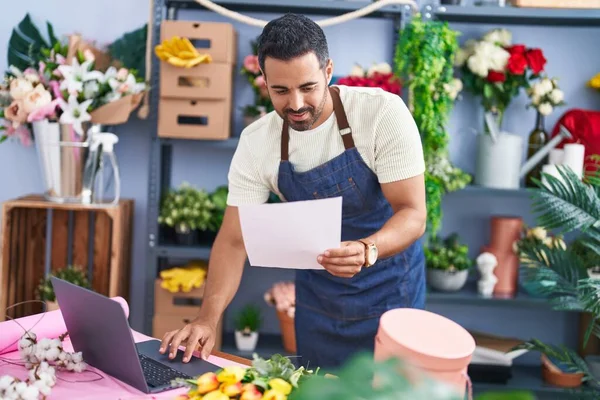 Young hispanic man florist using laptop reading paper at florist