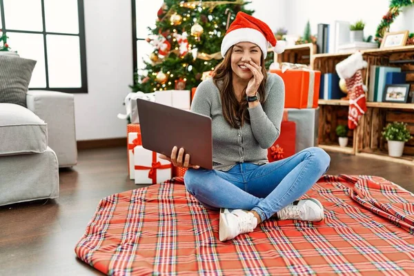 若いですラテン女性使用してノートパソコンで座ってクリスマスツリー笑いと恥ずかしいクスクスカバー口で手 ゴシップやスキャンダルの概念 — ストック写真