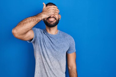 Mavi arka planda duran sakallı bir Orta Doğulu. Gülümsüyor ve elleri yüzünde, şaşırtmak için gözleri örtüyor. kör kavram. 