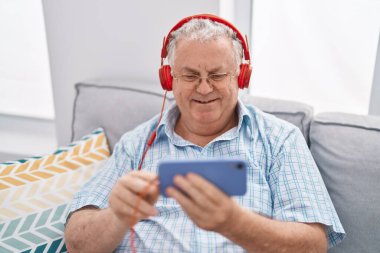Orta yaşlı, kır saçlı bir adam akıllı telefondan video izliyor. Evdeki kanepede oturuyor.
