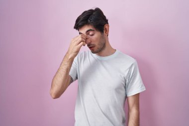 Young hispanic man standing over pink background tired rubbing nose and eyes feeling fatigue and headache. stress and frustration concept. 