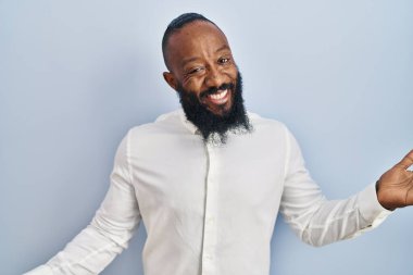 Mavi arka planda duran Afro-Amerikan adam gülümseyerek gülümsüyor. Dostça bir hoş geldin, olumlu ve kendinden emin selamlar. 