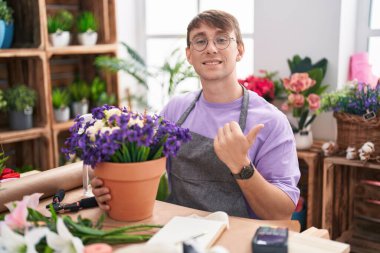 Caucasian blond man working at florist shop pointing to the back behind with hand and thumbs up, smiling confident 