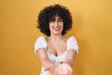 Sarı arka planda kıvırcık saçlı, elleri avuçlarında gülümseyen ve el hareketi yapan genç esmer kadın. Bekleme ve koruma 