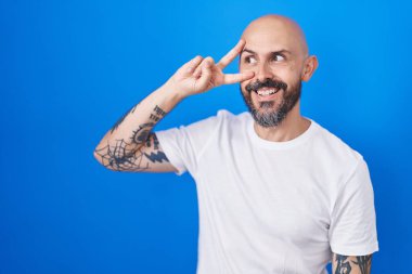 Mavi arka planda dikilen İspanyol dövmeli adam barış sembolü yapıyor. Yüzünde parmakları var, gülümsüyor ve zaferini gösteriyor. 