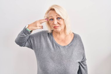 Orta yaşlı beyaz bir kadın beyaz arka planda durup ateş ediyor ve kendini ve parmaklarını silah gibi kullanıyor, intihar jesti yapıyor.. 