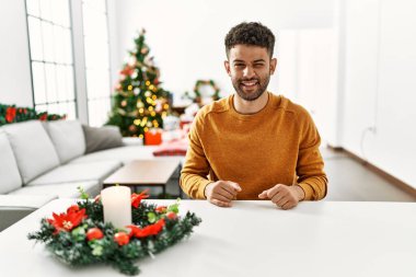 Arap genç adam Noel ağacının yanında oturmuş kameraya seksi bir ifadeyle bakıyor, neşeli ve mutlu bir yüzle.. 