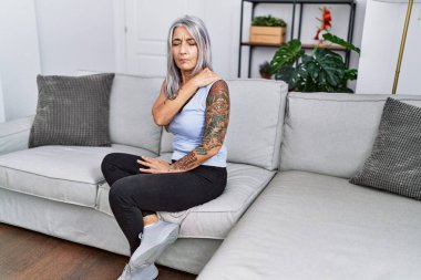 Orta yaşlı, kır saçlı, boynu ağrıyan bir kadın evdeki kanepede oturuyor.