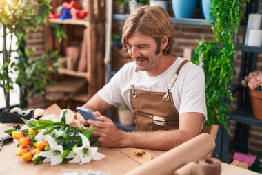Çiçekçide akıllı telefon kullanarak gülümseyen genç sarışın adam.