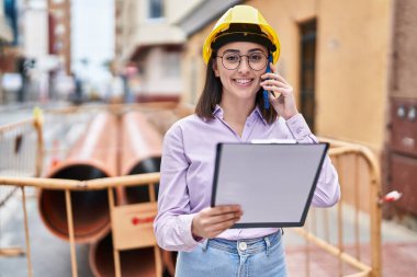 Young hispanic woman architect talking on the smartphone reading document at street