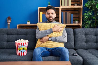 Hispanic man with beard eating popcorn watching a movie at home skeptic and nervous, frowning upset because of problem. negative person. 