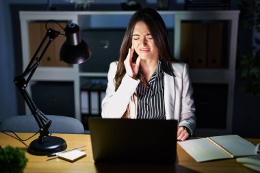 Geceleri ofiste çalışan esmer bir kadın diş ağrısı ya da diş hastalığından dolayı elleri ve elleri ile dudaklarına dokunan bir dizüstü bilgisayarla çalışıyor. dişçi kavramı. 