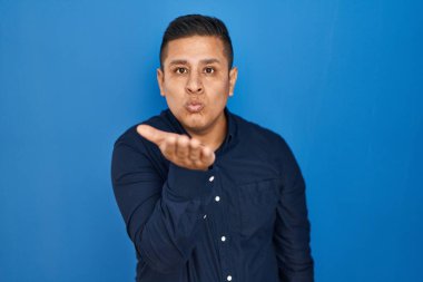 Latin asıllı genç bir adam mavi arka planda durup kameraya bakarak havada bir öpücük savuruyor ve sevimli ve seksi oluyor. aşk ifadesi. 