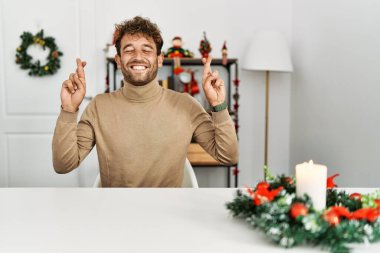 Noel süslemesi masasında oturan sakallı yakışıklı genç adam umut ve gözler kapalı gülümseyerek el işareti yapıyor. Şans ve batıl inançlar. 