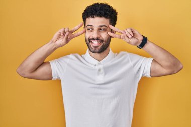 Sarı arka planda duran Arap adam barış sembolü yapıyor. Yüzünde parmaklar, gülümseyerek zaferi gösteriyor. 