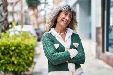 Orta yaşlı kadın gülümsüyor. Kollarını kavuşturup caddede duruyor.