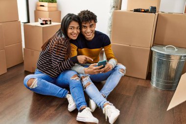 Genç Latin çift yeni evlerinde yerde otururken akıllı telefon kullanıyor..