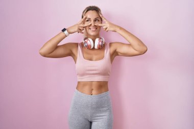Young blonde woman wearing sportswear and headphones doing peace symbol with fingers over face, smiling cheerful showing victory 